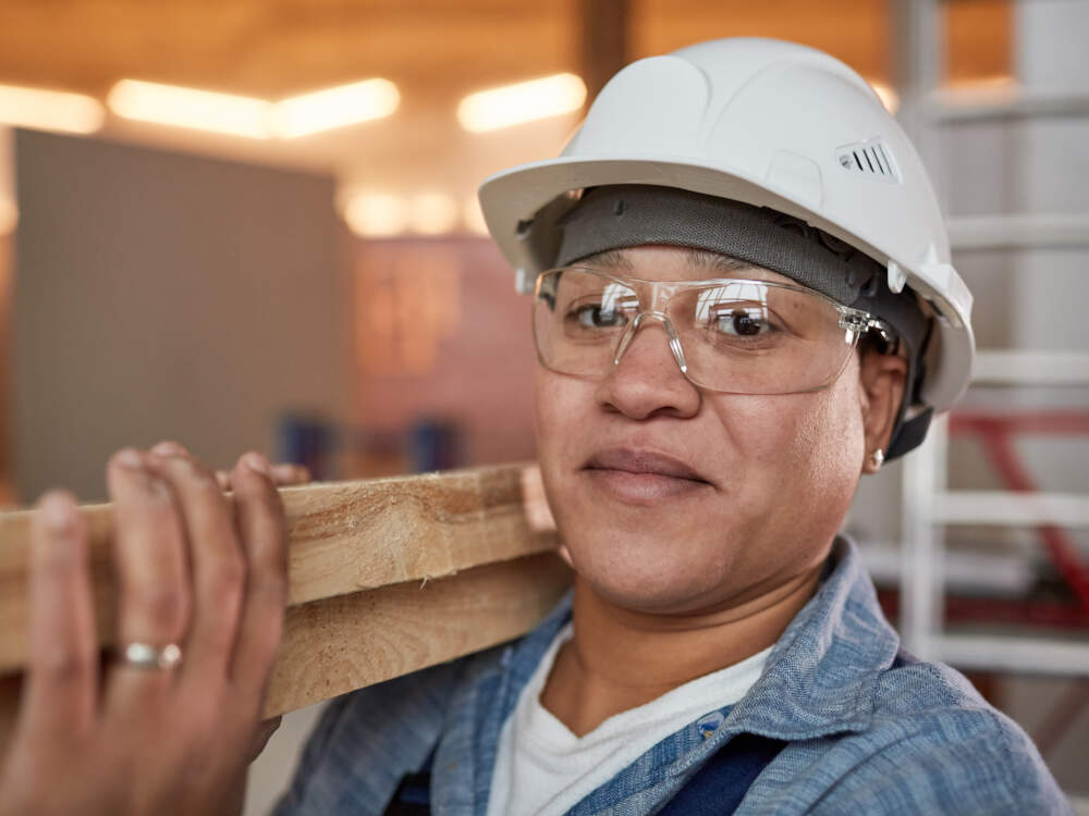 A cheerful female construction worker carries lumber with a hand featuring a wedding ring showcasing our commitment to fair wages and supportive communities in Muskoka, Ontario.