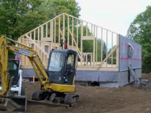 A carpenter plumbs a gable end wall using a level in the background behind a rented hydraulic excavator.