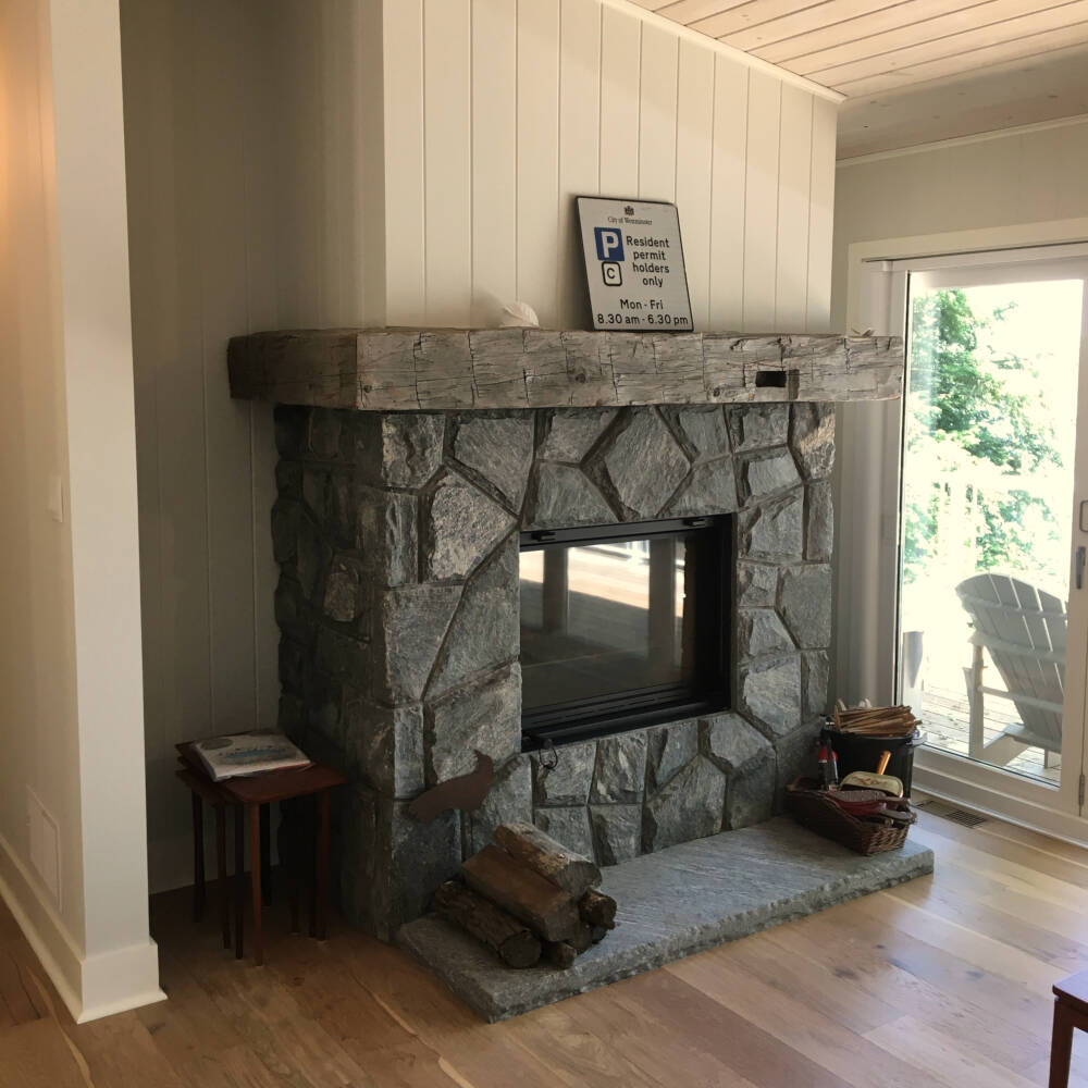Woodburning masonry fireplace with rustic reclaimed barn timber mantel in a sunlit Living Room overlooking Lake Muskoka.