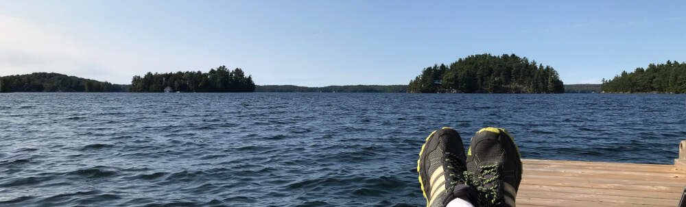 A sunbathers toes are visible on the end of a dock sanwiched between blue lake water, forested shoreline and blue summer sky.