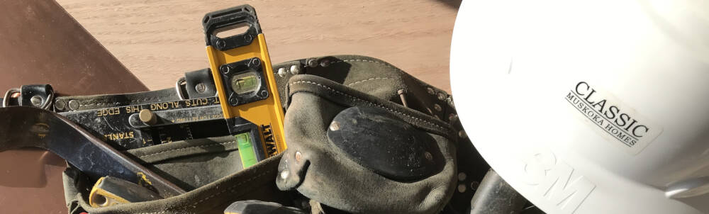 A black carpenters belt with pry bar, square and level are pictured beside a Classic Muskoka Homes branded hard hat to refrence construction jobs in Muskoka, Ontario.